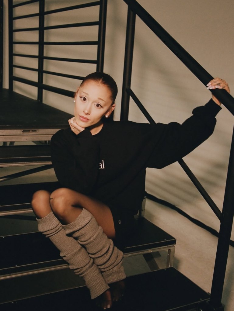 Ariana Grande in a black sweatshirt, black shorts, and gray leg warmers, sitting on a set of stairs.