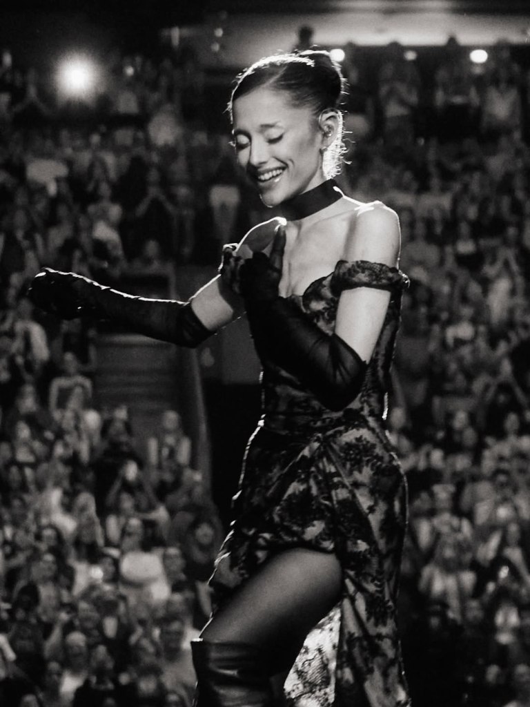 Black and white photo of Ariana Grande performing on stage, smiling with her eyes closed.