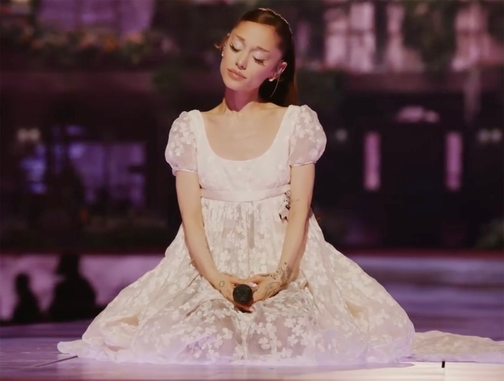 Ariana Grande wearing a white floral dress, sitting on stage with a microphone in her hands.