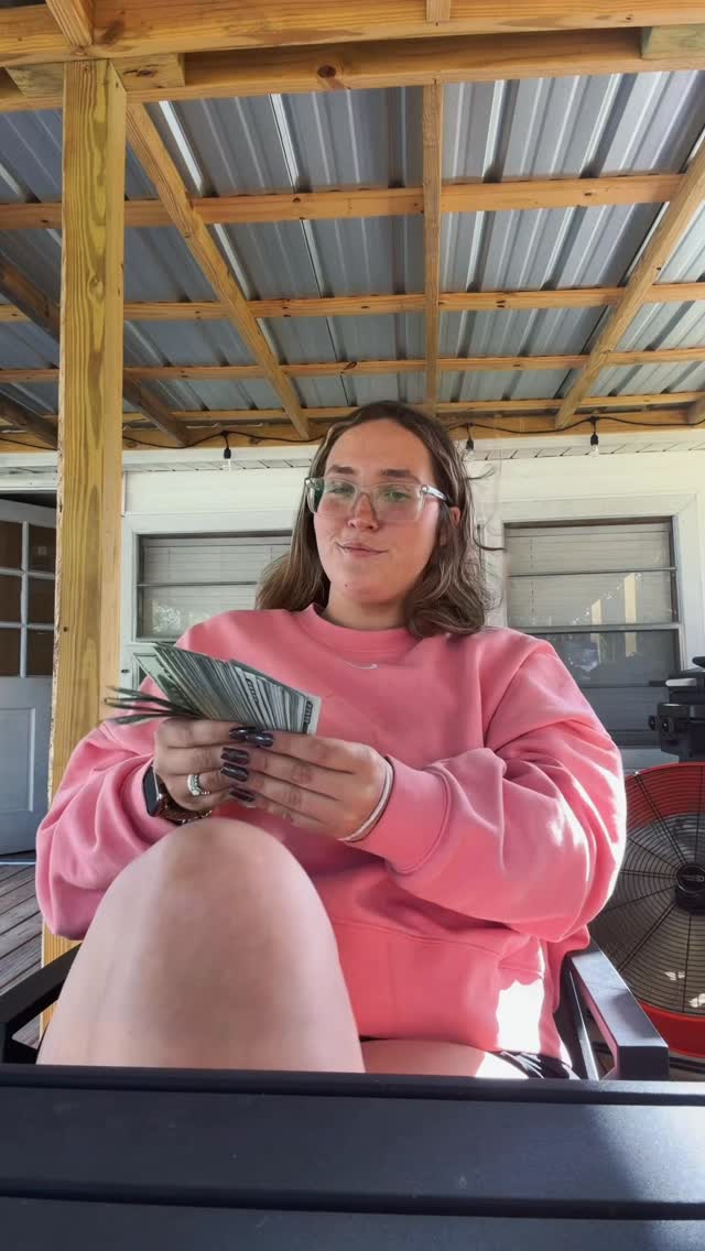 A woman in a pink sweatshirt sitting on a patio counting a stack of cash.