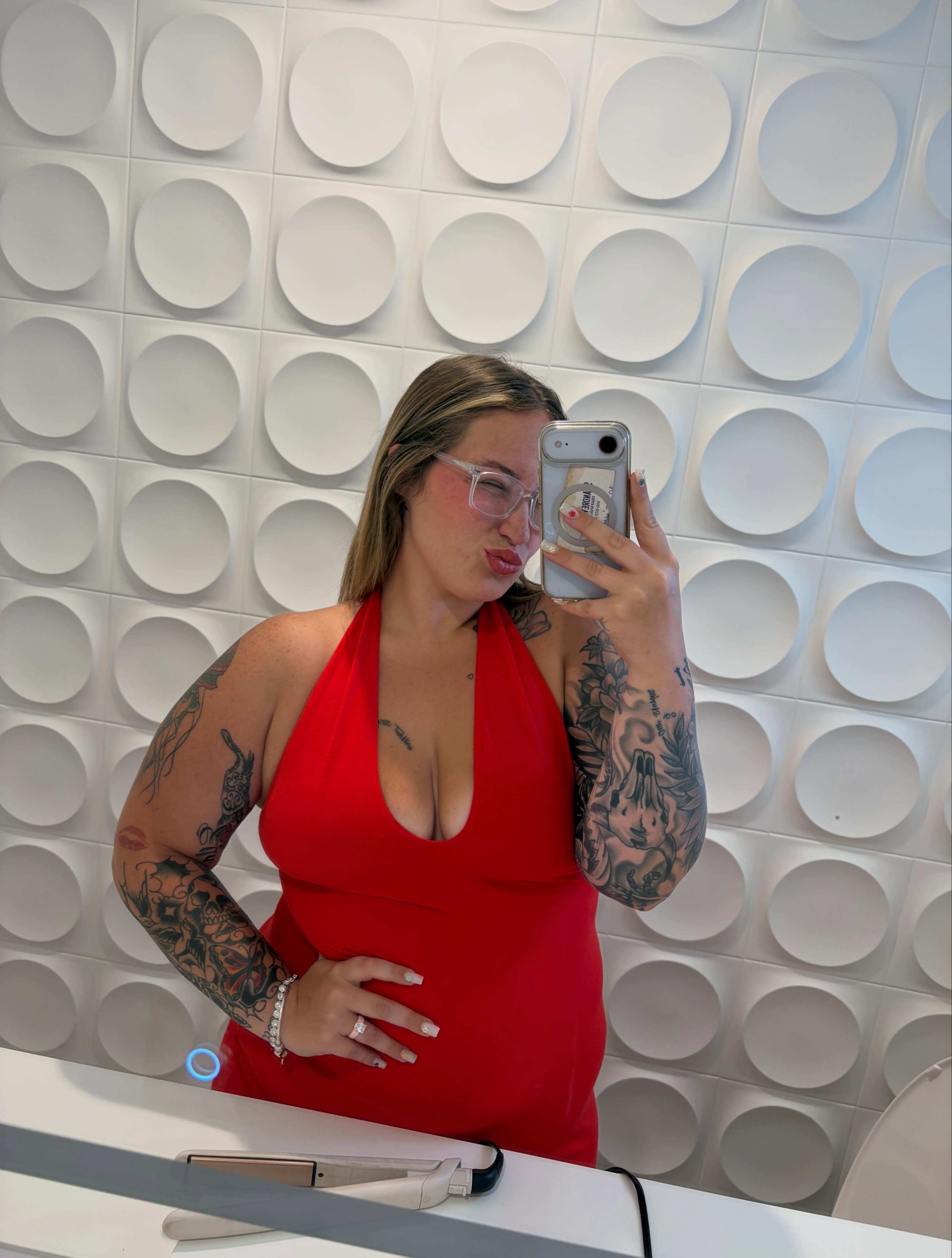 A woman with long blonde hair and glasses takes a mirror selfie, posing in a red halter top and showing off her tattooed arms.