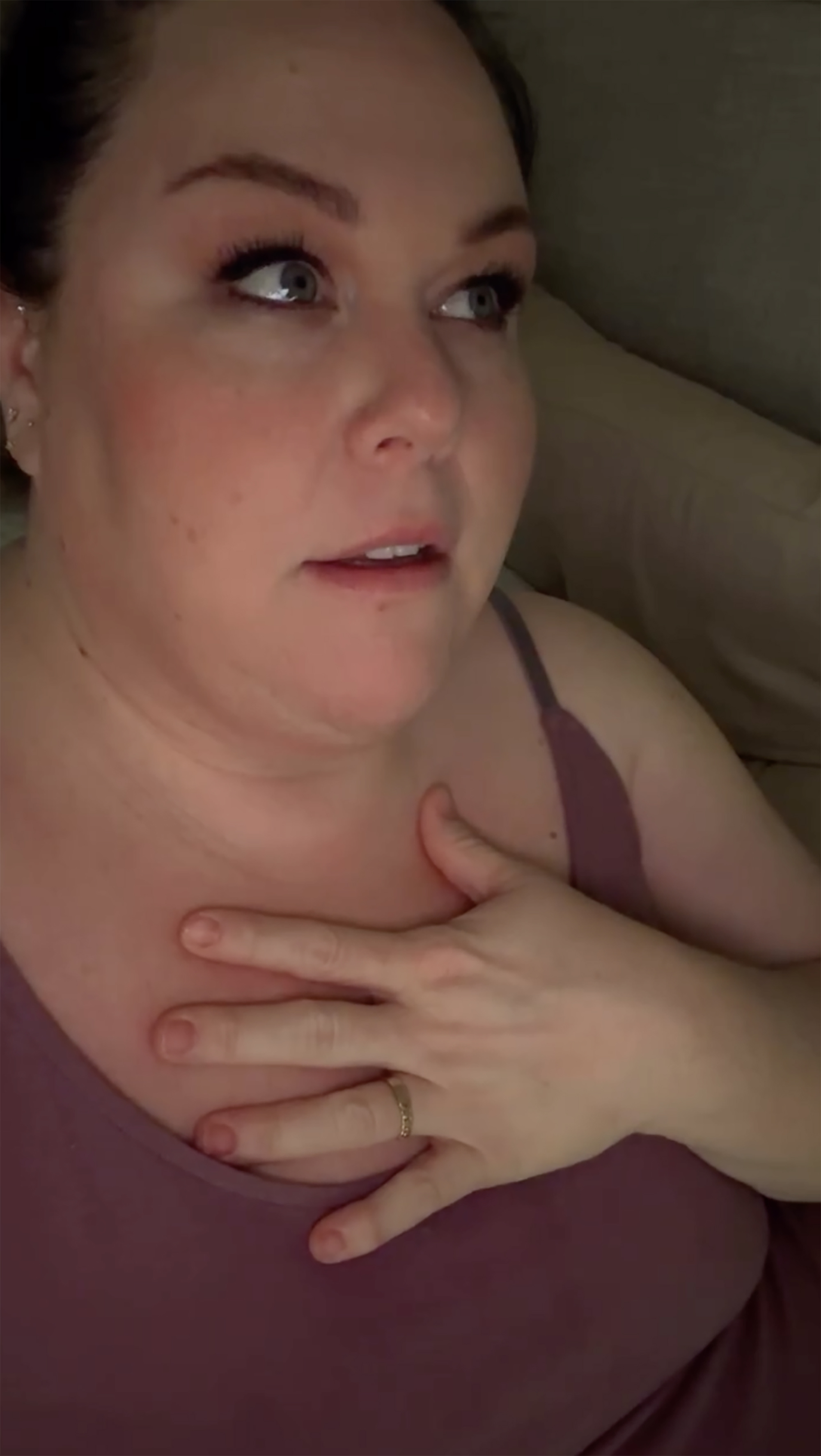 Close-up of a person with light skin and dark hair in a burgundy top, holding a hand to their chest, with a gold ring on their ring finger, looking to the side with their mouth slightly open.