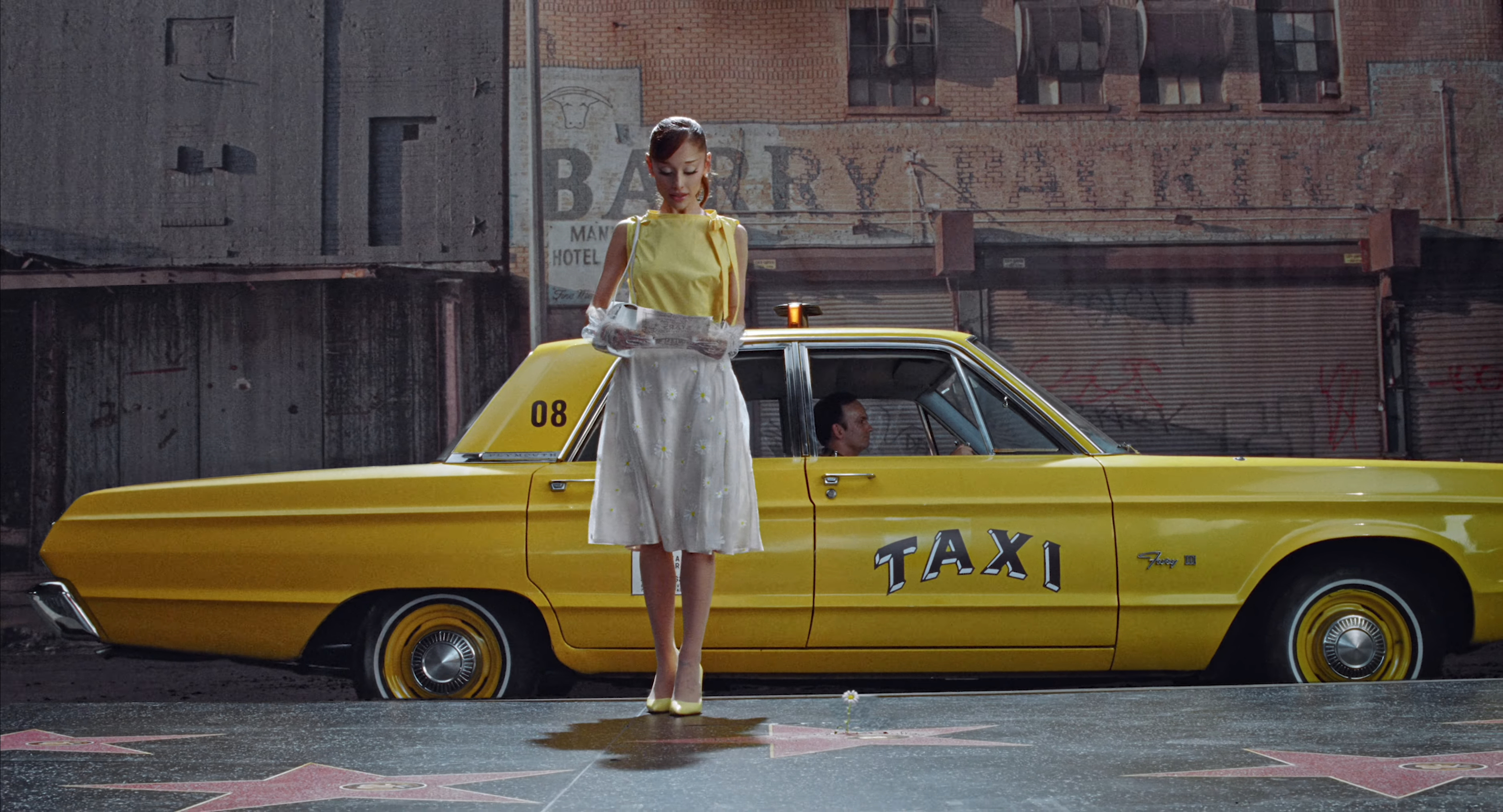 Ariana Grande in a yellow top and daisy-embroidered skirt stands in front of a yellow taxi, looking at a map.