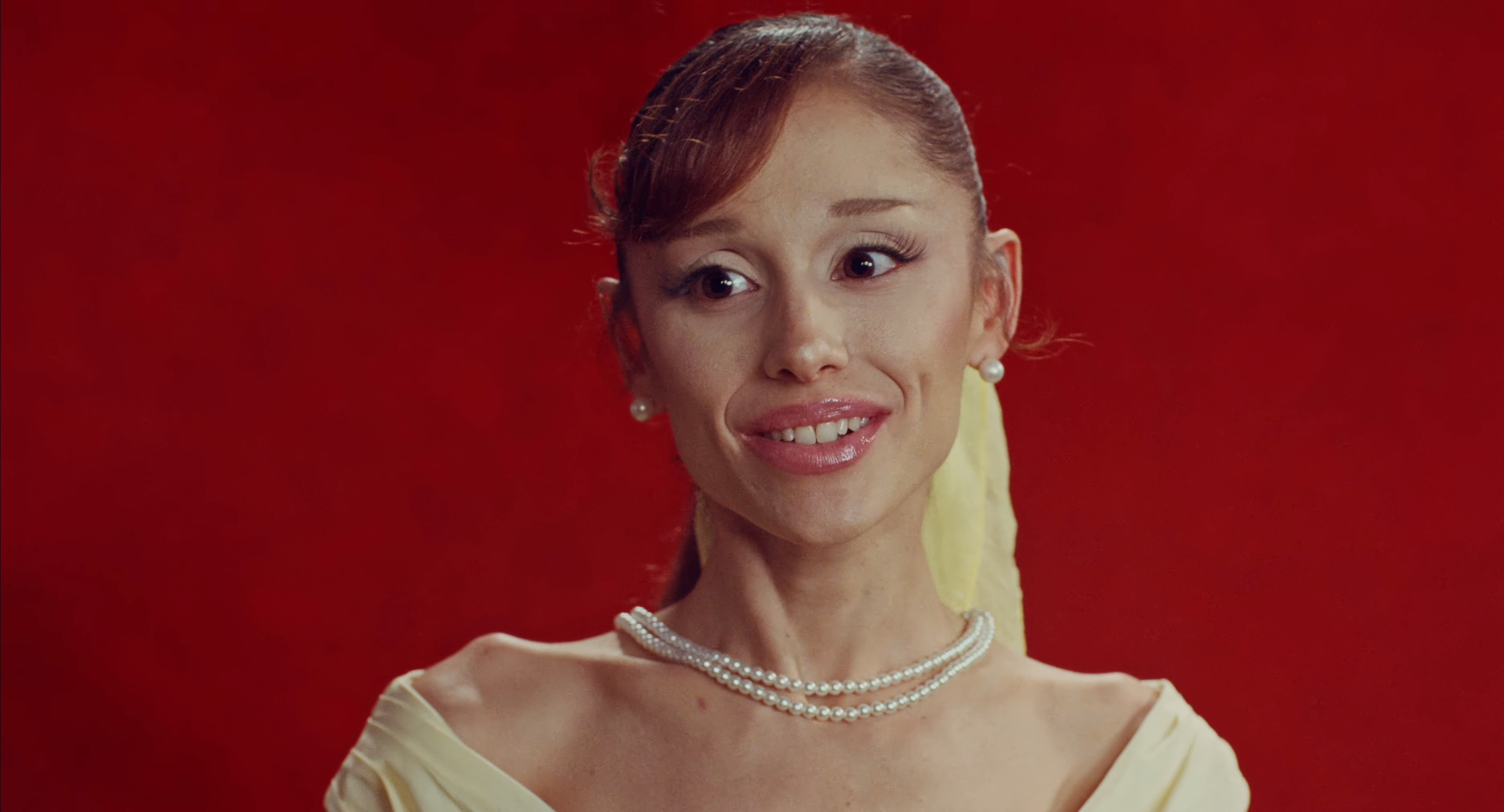 Ariana Grande smiles against a red background, wearing a pearl necklace and earrings.