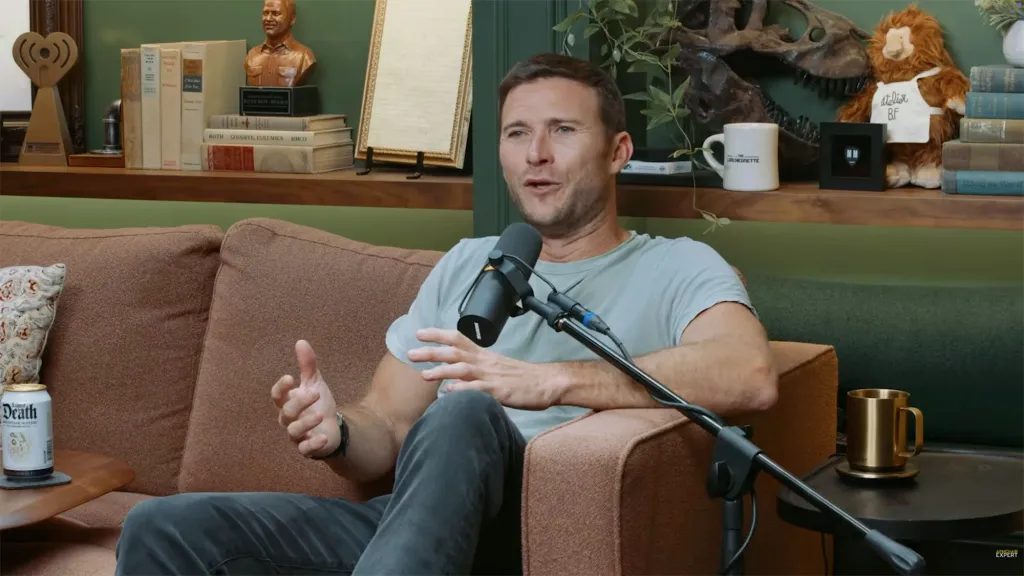 Scott Eastwood speaking into a microphone while sitting on a couch.