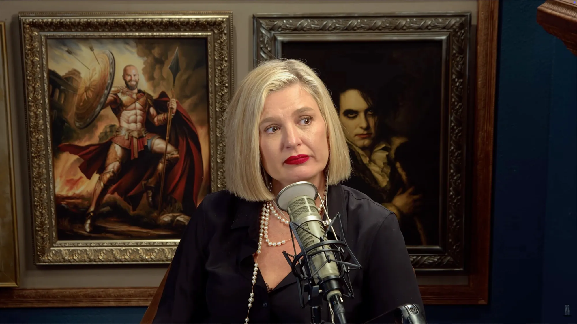 Christina Pazsitzky with a microphone in front of two paintings, one of a Spartan warrior and another of a person with dark eye makeup.