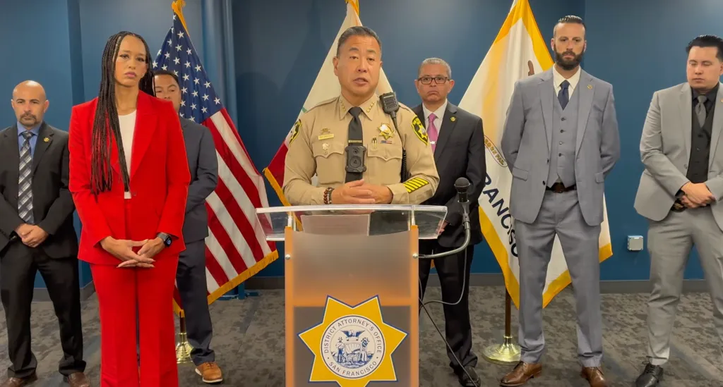 San Francisco Sheriff Paul Miyamoto speaking at a podium with several individuals standing behind him.