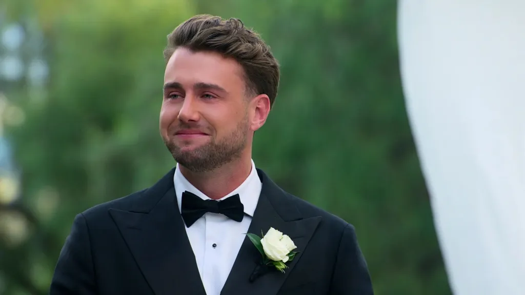 A groom in a tuxedo with a white boutonnière smiles and looks emotional.