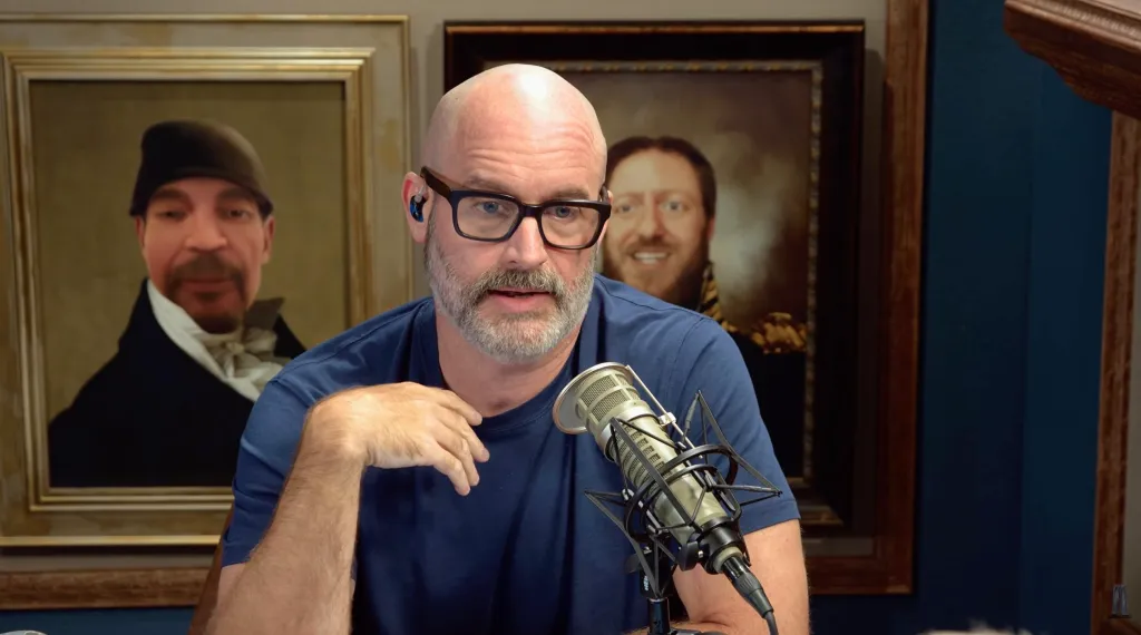 A man with a bald head, glasses, and a gray beard speaking into a microphone, with two portraits in the background featuring faces superimposed onto historical figures.
