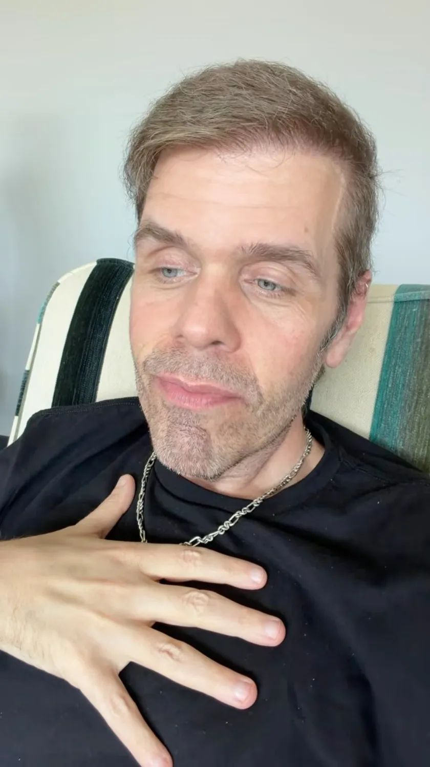 A man with blue eyes and light brown hair in a black shirt with a silver chain rests his left hand over his heart.