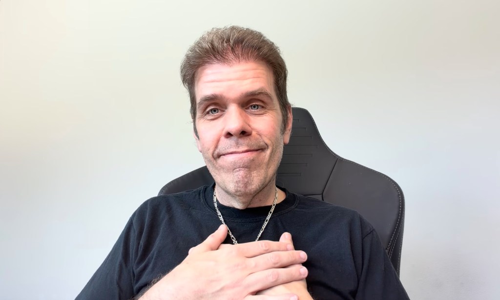 A man in a black shirt and silver chain sits in a gaming chair with his hands over his heart.