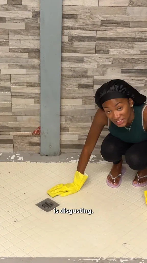 Big Brother Taylor cleaning a shower, wearing a black shower cap, green top, black pants, and light purple flip-flops, while scrubbing with a yellow gloved hand and sponge.