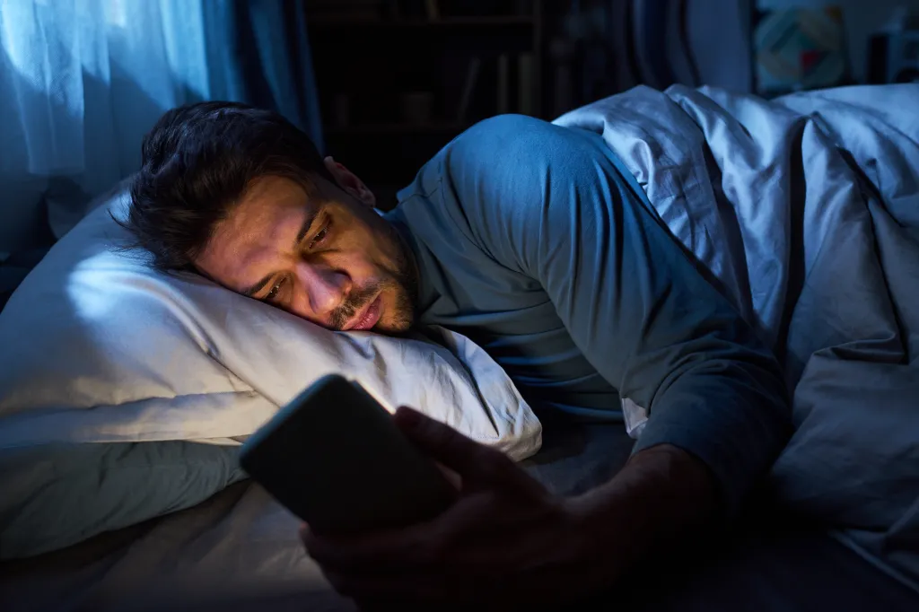 A young man lies in bed, illuminated by the light of his smartphone, late at night.
